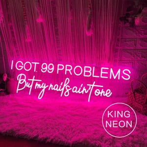 K&ouml;nnte beinhalten: Ein pinkfarbenes Neon-Schild mit dem Text "I got 99 problems but my nails ain't one". Das Schild leuchtet und h&auml;ngt an einer Wand mit einem wei&szlig;en und rosa Hintergrund. Das Schild wird von King Neon hergestellt.