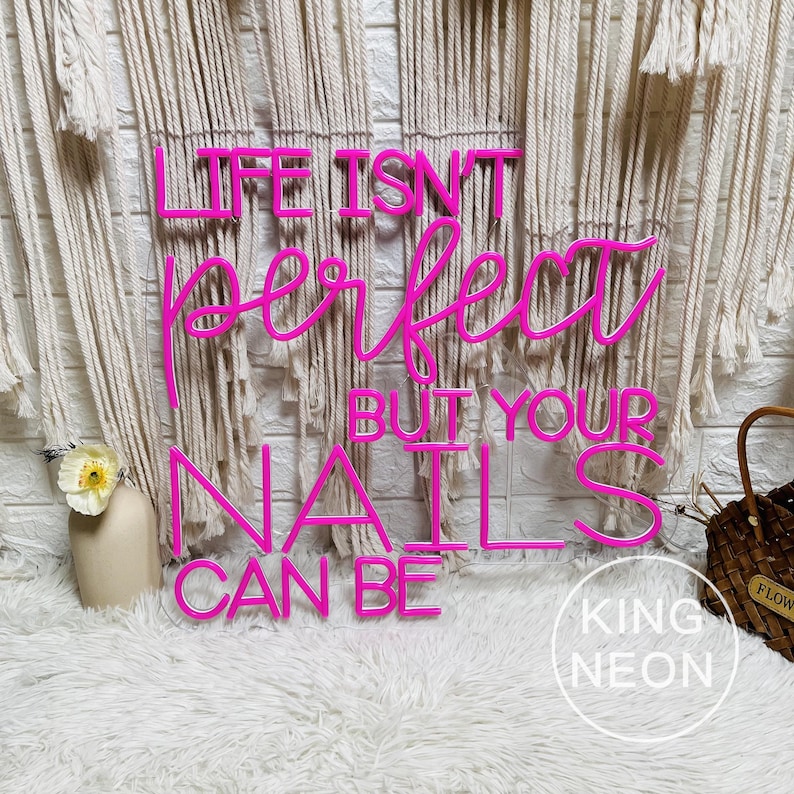 Op de afbeelding: Een felroze neonbord met de tekst "LIFE ISN'T PERFECT BUT YOUR NAILS CAN BE." Het bord staat tegen een achtergrond van macram&eacute; wandkleden en een zacht wit tapijt. Het King Neon logo is zichtbaar.