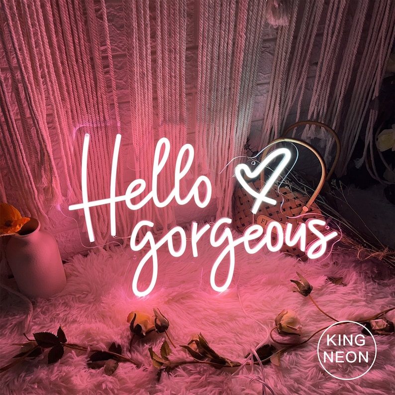 K&ouml;nnte beinhalten: Rosa Neon-Schild mit dem Text "Hello gorgeous" und einem Herz. Das Schild steht auf einer wei&szlig;en, flauschigen Oberfl&auml;che mit Blumen und einem Weidenkorb.