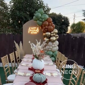 May include: A festive Thanksgiving table setting with a pink tablecloth, gold chairs, and a white pumpkin centerpiece. A balloon arch in shades of green, brown, and gold frames a sign that reads "Happy Friendsgiving."