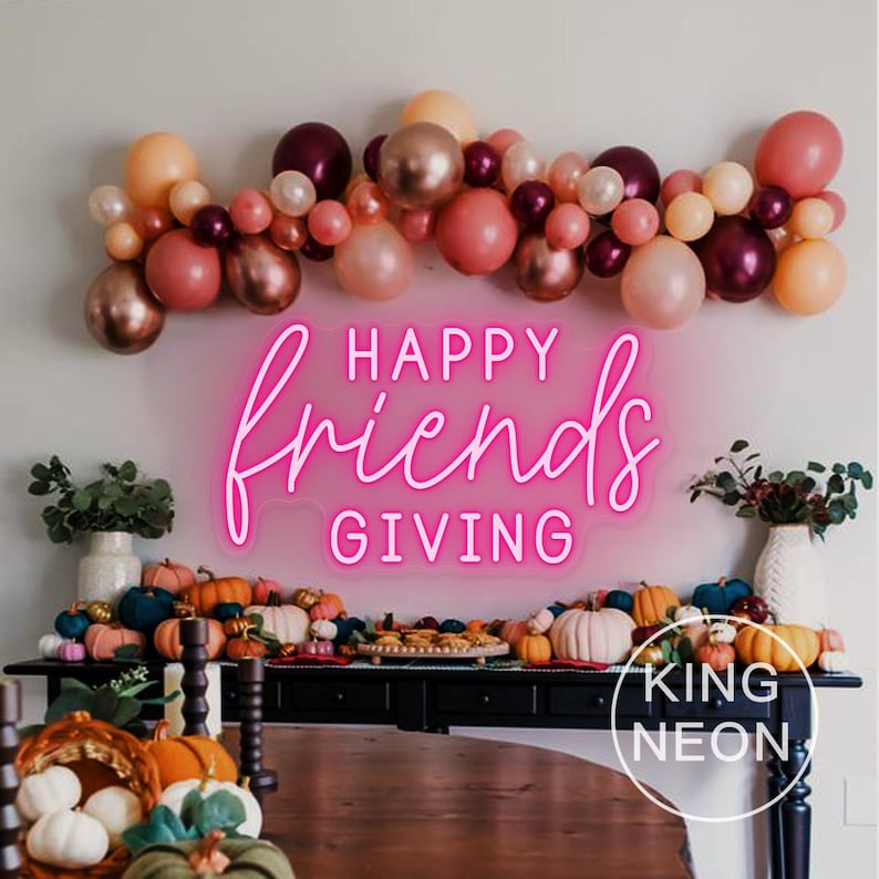 May include: A festive Thanksgiving display featuring a neon sign that reads "Happy Friends Giving" in pink. The sign is above a table decorated with pumpkins, gourds, and a balloon garland in shades of pink, peach, and burgundy. The King Neon logo is visible.