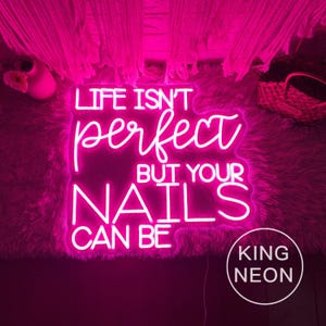 Op de afbeelding: Een helder roze neonbord met de tekst "LIFE ISN'T perfect BUT YOUR NAILS CAN BE." Het bord staat tegen een pluizige roze achtergrond en bevat het "KING NEON" logo.