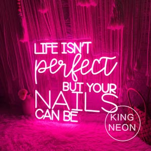Op de afbeelding: Een helder roze neonbord met de tekst "LIFE ISN'T perfect BUT YOUR NAILS CAN BE." Het bord wordt omgeven door een roze, zacht oppervlak en een decoratieve achtergrond. Het bord bevat ook de tekst "KING NEON" in een cirkel.