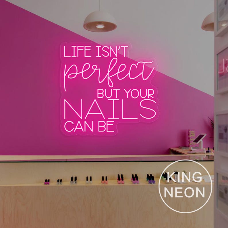 Op de afbeelding: Een neonbord met de tekst "LIFE ISN'T perfect BUT YOUR NAILS CAN BE" in witte letters tegen een felroze muur. De afbeelding toont ook een display van nagellak in een nagelsalon.