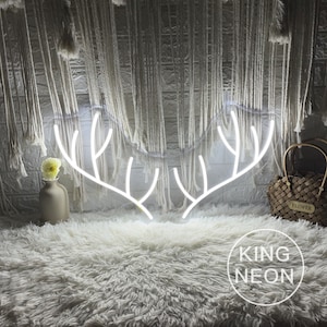 Può includere: Un cartello al neon bianco a forma di corna di cervo. Il cartello è illuminato e si trova su un tappeto bianco e soffice. Il testo "KING NEON" è visibile nell'angolo in basso a destra dell'immagine.
