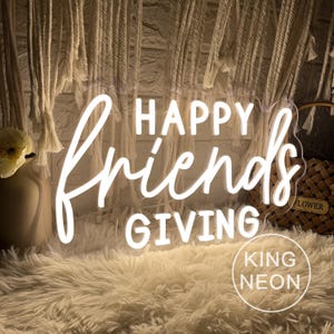 May include: A white neon sign with the words "HAPPY Friends GIVING" in a cursive font. The sign is on a fluffy white surface, with a woven basket and decorative wall hangings in the background. The sign also has the text "KING NEON".
