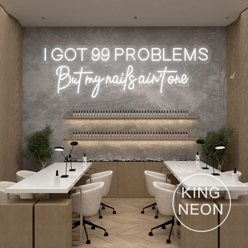 K&ouml;nnte beinhalten: Ein wei&szlig;es Neon-Schild mit dem Text "I got 99 problems but my nails ain't one". Das Schild ist an einer grauen Wand in einem Nagelstudio angebracht. Im Vordergrund stehen zwei wei&szlig;e Manik&uuml;retische mit schwarzen St&uuml;hlen.