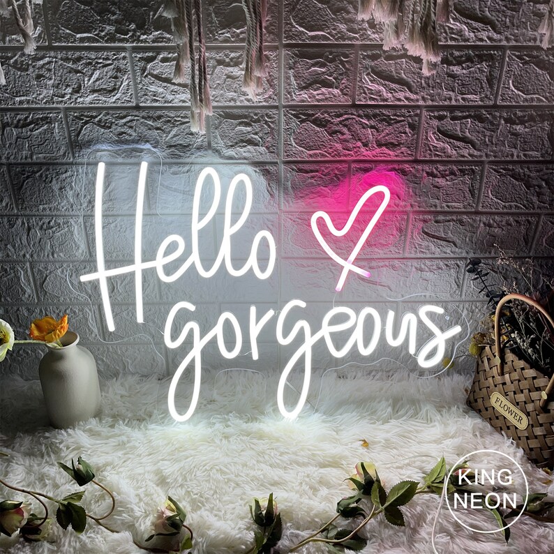 K&ouml;nnte beinhalten: Ein wei&szlig;es Neon-Schild mit dem Text "Hello gorgeous" und einem rosa Herzen. Das Schild ist an einer Backsteinwand montiert und leuchtet. Das Schild wird von King Neon hergestellt.