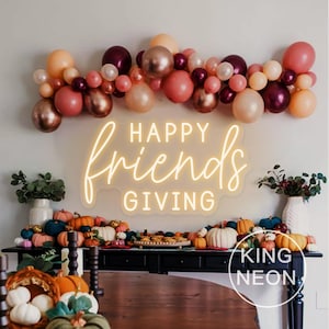 May include: A festive Thanksgiving display featuring a neon sign that reads "HAPPY friends GIVING" in a cursive font. The sign is illuminated with a warm glow, complemented by a balloon garland in shades of pink, peach, and burgundy. Pumpkins and gourds decorate the table.