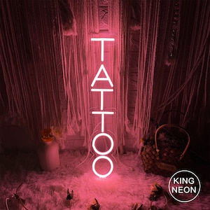 May include: Pink neon sign with the word "TATTOO" in white letters. The sign is lit up and is sitting on a white fluffy surface.