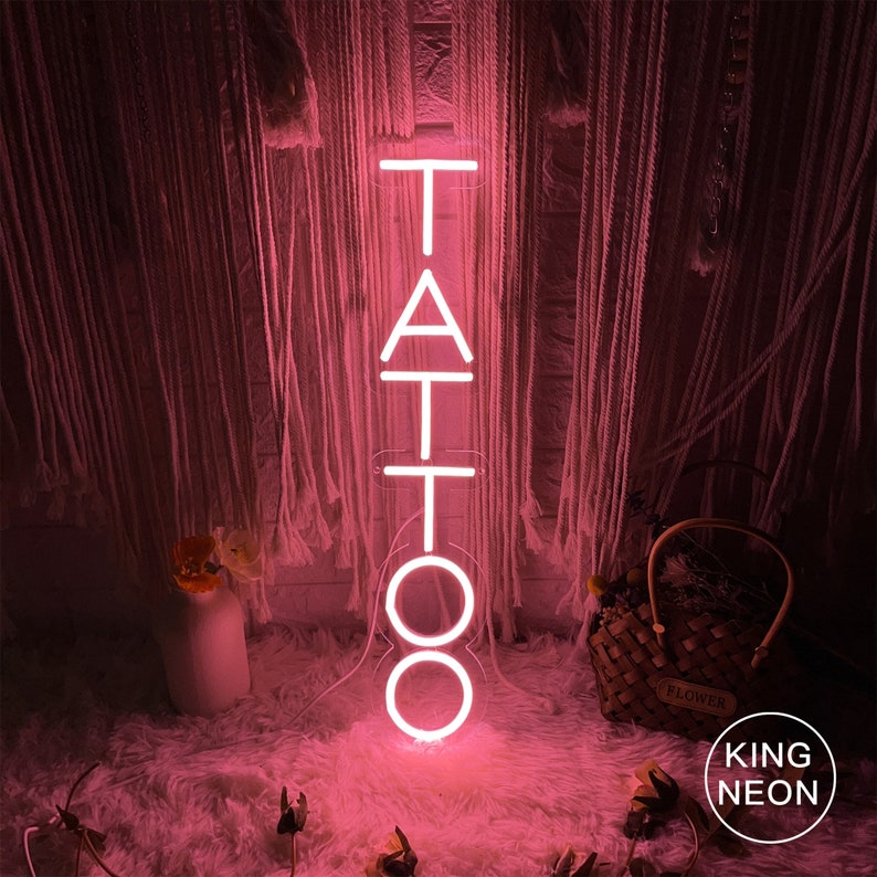 May include: Pink neon sign with the word "TATTOO" in white letters. The sign is lit up and is sitting on a white fluffy surface.