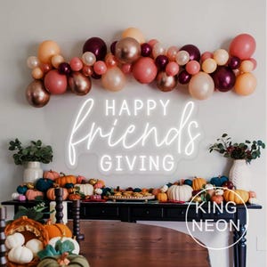 May include: A festive Thanksgiving display featuring a neon sign that reads "Happy Friends Giving". The scene includes a balloon garland in shades of pink, peach, and burgundy, and a table adorned with pumpkins and gourds.