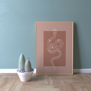 May include: A framed print with a minimalist design of a white snake with celestial symbols on a dusty rose background.