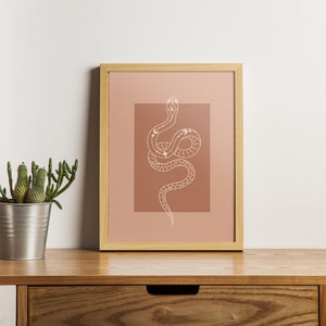 May include: A framed print of a line drawing of a snake with celestial bodies on its body. The snake is coiled and has its head raised. The background is a muted terracotta color.