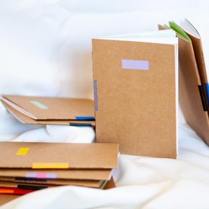 Könnte beinhalten: Ein Stapel brauner Notizbücher mit bunten Washi-Tape-Akzenten. Die Notizbücher liegen auf einer weißen Stoffoberfläche.
