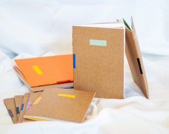 Puede incluir: Una pila de cuadernos marrones con acentos de cinta de colores. Los cuadernos tienen cubiertas en blanco y son perfectos para llevar un diario, tomar notas o dibujar.