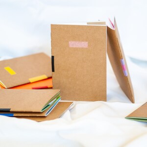 Könnte beinhalten: Ein Stapel brauner Notizbücher mit bunten Klebebandakzenten. Die Notizbücher sind aus Recyclingpapier hergestellt und haben ein natürliches, rustikales Aussehen.