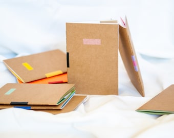 Puede incluir: Una pila de cuadernos marrones con detalles de cinta de colores. Los cuadernos están hechos de papel reciclado y tienen un aspecto natural y rústico.