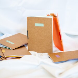 Könnte beinhalten: Ein Stapel brauner Notizbücher mit bunten Klebebandakzenten. Die Notizbücher liegen auf einer weißen Oberfläche.