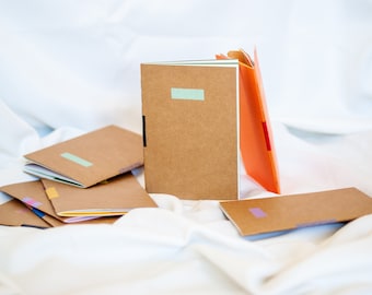 Puede incluir: Una pila de cuadernos marrones con detalles de cinta de colores. Los cuadernos están sobre una superficie blanca.