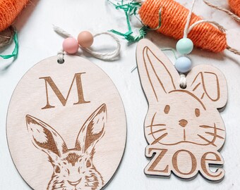 May include: Two wooden Easter ornaments. One is egg-shaped with a rabbit engraving and the letter "M". The other is a bunny head with the name "Zoe". Both have pastel-colored beads and twine. Orange carrot decorations are in the background.