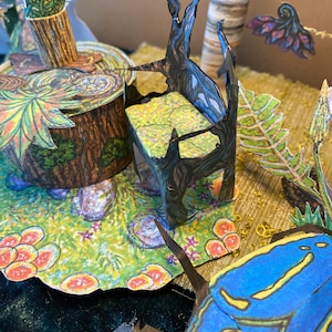 Könnte beinhalten: Eine Miniatur-Papierbastel-Szene mit einem schwarzen Baumstumpf-Stuhl, einem grün-braunen Baumstumpf-Tisch und einem blauen und gelben Papierinsekt.