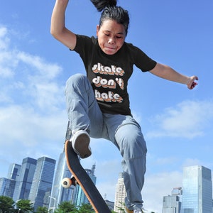 Puede incluir: Una persona que lleva una camiseta negra con el texto "skate don't hate" en colores arcoíris, está saltando en el aire mientras monta en patineta. La persona lleva pantalones vaqueros azules y zapatillas amarillas. El fondo es un horizonte urbano.