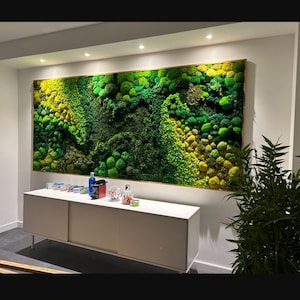 May include: A green moss wall art piece hangs on a white wall above a white cabinet with a wood top. The moss wall art is framed in a light brown wood frame.