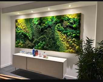 May include: A green moss wall art piece hangs on a white wall above a white cabinet with a wood top. The moss wall art is framed in a light brown wood frame.