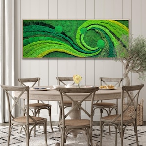 May include: A framed moss wall art piece with a swirling green and yellow design. The art is hanging on a white wall above a wooden dining table with chairs.