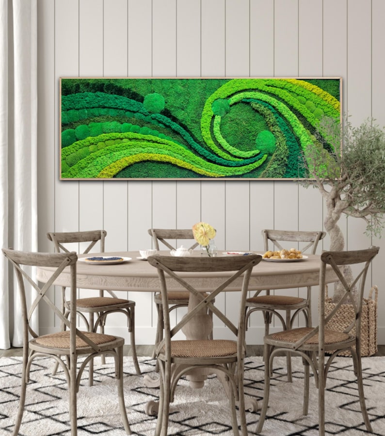 May include: A framed moss wall art piece with a swirling green and yellow design. The art is hanging on a white wall above a wooden dining table with chairs.