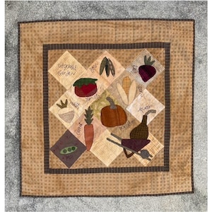 May include: A quilted square featuring a vegetable garden theme. The design includes appliqued images of vegetables like tomatoes, carrots, and pumpkins, with the words "Vegetable Garden" and the names of the vegetables. The quilt has a brown and tan color scheme.