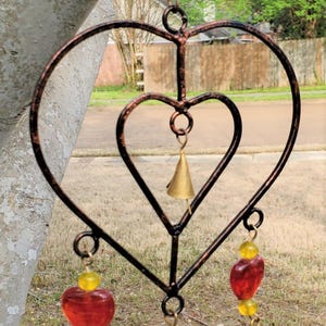 May include: A heart-shaped metal wind chime with red and yellow glass beads and gold bells. The chime is hanging from a tree branch.