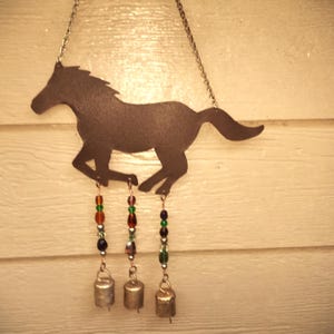 May include: A metal horse silhouette wind chime. The dark brown horse is in a running pose, suspended by a chain. Colorful beads in green, orange, and blue hang below, leading to small silver bells. The background is a white wooden surface.
