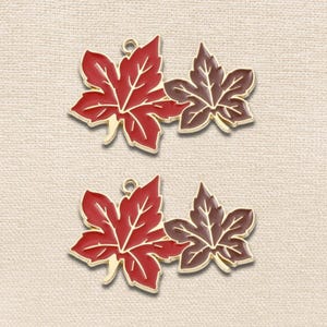 May include: Two identical metal charms, each featuring a pair of connected maple leaves. One leaf is filled with bright red enamel, while the smaller adjacent leaf is filled with dark brown enamel. Both leaves have gold-toned metal outlines.
