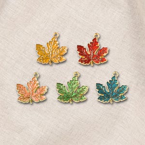 May include: Five metal maple leaf charms featuring gold-toned outlines and enamel fill in yellow, red, light orange, green, and teal. Each leaf-shaped pendant includes a small circular loop at the top for attachment, showcasing detailed vein textures.