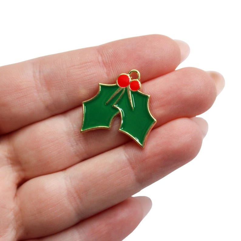 May include: A small metal charm shaped like two holly leaves with two bright red berries at the top. The leaves are filled with glossy green enamel, and the entire piece is outlined in a thin gold-colored metal border with a small loop at the top.