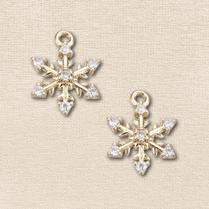 May include: Two gold-toned snowflake charms featuring a central clear stone and additional clear stones set at the end of each of the six points. Each charm includes a small circular jump ring at the top for attachment to jewelry or accessories.