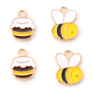 May include: Four gold-colored metal charms with a bee theme. Two charms feature a bee with black and yellow stripes, white wings, and a gold loop. The other two charms feature a honey pot with black and yellow stripes, a white lid, and a gold loop.