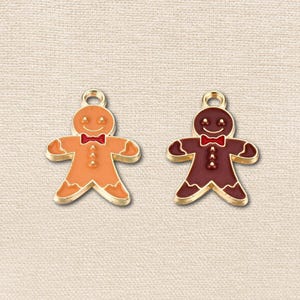 May include: Two gingerbread man charms with gold-tone metal outlines and enamel details. One charm is light orange and the other is dark brown. Both feature a red bow tie, white icing accents on the limbs, and a small loop at the top for hanging.