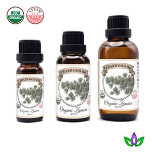 Op de afbeelding: Drie bruine glazen flessen cederhoutolie met zwarte doppen. Elke fles heeft een etiket met de tekst "CEDARWOOD OIL" en een botanische illustratie. De etiketten bevatten ook "Organic Zamara" en het volume: 15 ml, 30 ml en 60 ml.