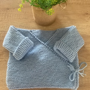 Op de afbeelding: Een lichtblauwe gebreide babywikkel met opgevouwen mouwen en striksluitingen. De wikkel is gemaakt van een zacht, getextureerd garen en is ontworpen om een baby warm te houden. De wikkel wordt getoond op een houten oppervlak.
