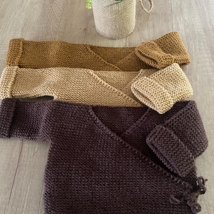 Op de afbeelding: Drie gebreide babywikkelvesten in bruin-, beige- en paarse tinten. Elk vest heeft lange mouwen en een wikkelontwerp. Een kleine plant in een pot staat op de achtergrond.