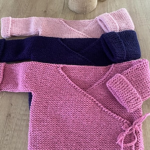 Puede incluir: Se muestran tres cárdigans de bebé de punto en tonos rosa y azul marino. Los cárdigans tienen un diseño envolvente con mangas largas y cierres de lazo. Los cárdigans están hechos de un hilo suave y texturizado.