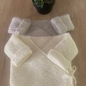 Puede incluir: Dos envolturas de bebé de punto, una blanca y otra gris claro, con mangas a juego. Las envolturas están hechas con un punto de tejido sencillo y tienen un cierre de nudo.