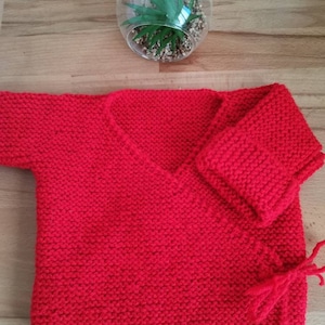 Puede incluir: Un cárdigan envolvente para bebé tejido a mano, de color rojo brillante. El diseño envolvente presenta un cuello en V y mangas largas. El cárdigan está hecho de un hilo suave y texturizado y tiene cierres de lazo. Un terrario decorativo de vidrio con una planta está en el fondo.