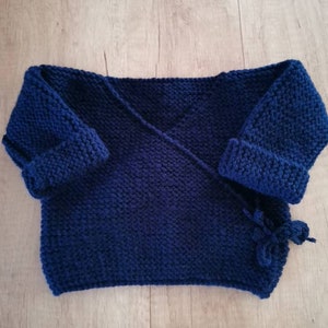 Op de afbeelding: Een donkerblauwe gebreide babytrui met een wikkel-stijl en een strik sluiting.