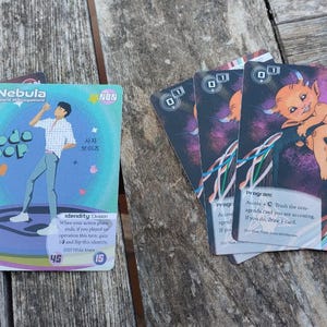 May include: A selection of trading cards with vibrant illustrations. One card displays a man in a white shirt and blue trousers with the words "Nebula" and "code pop". Other cards feature a cartoon devil.