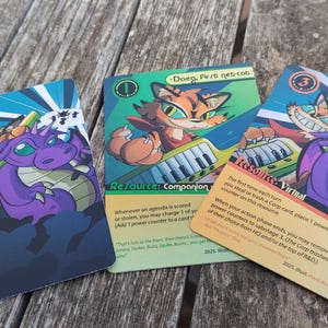 Pode incluir: Três cartas colecionáveis coloridas com ilustrações de fantasia. Uma carta apresenta um gato tocando um teclado, outra um dragão roxo e uma raposa, e a terceira um dragão e uma raposa. As cartas têm texto e estão sobre uma superfície de madeira.
