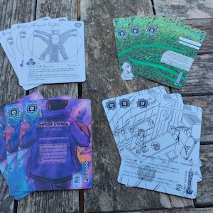 Puede incluir: Un conjunto de cartas de juego con diversas ilustraciones. Una pila presenta un diagrama de figura humana, otra un diseño verde, y otras representan a una persona con una chaqueta morada y una escena interior. Las cartas están dispuestas sobre una superficie de madera.
